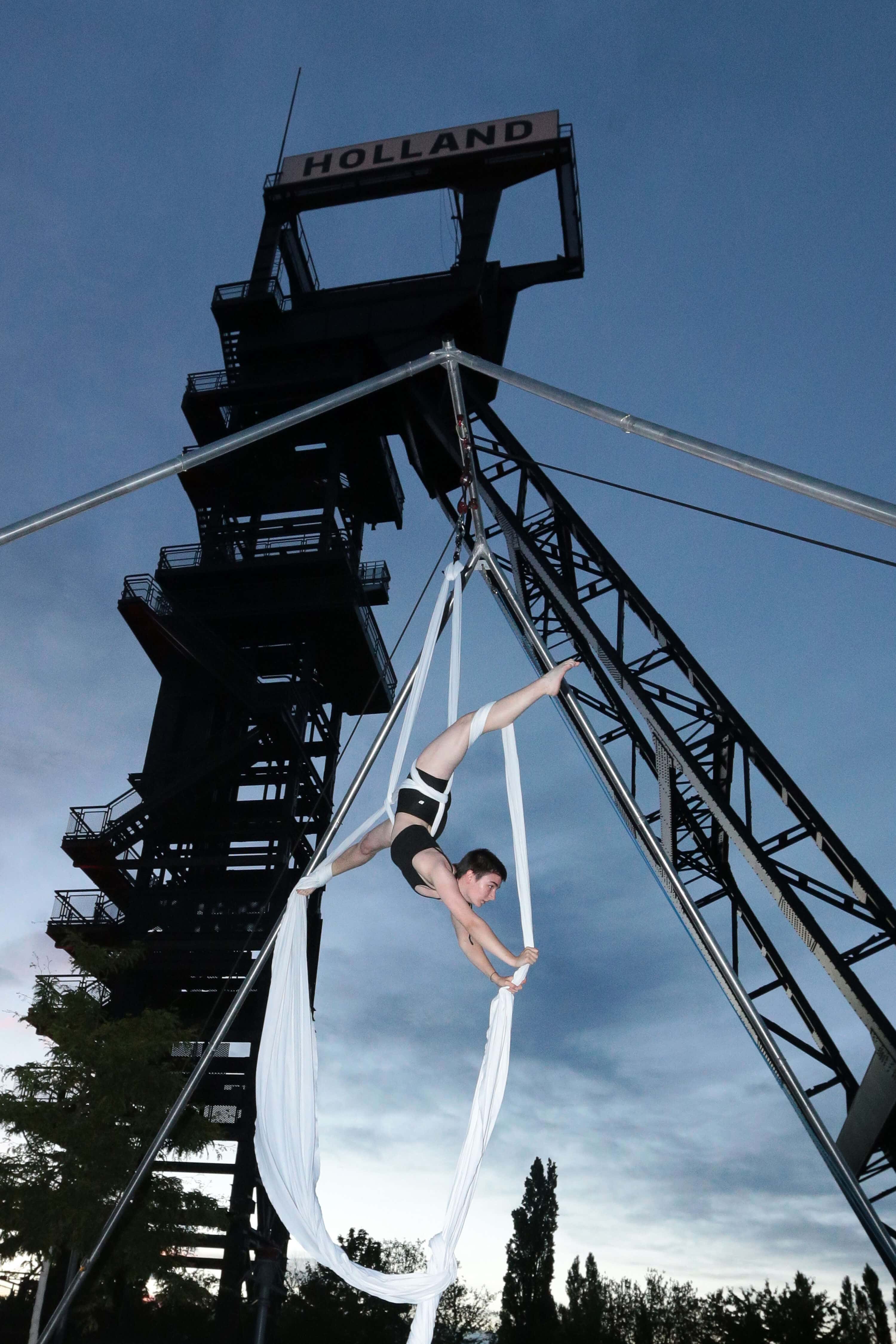 Luftartistin an weißen Tüchern vor dem illuminierten Fördergerüst der Zeche Holland in Bochum bei Abenddämmerung.