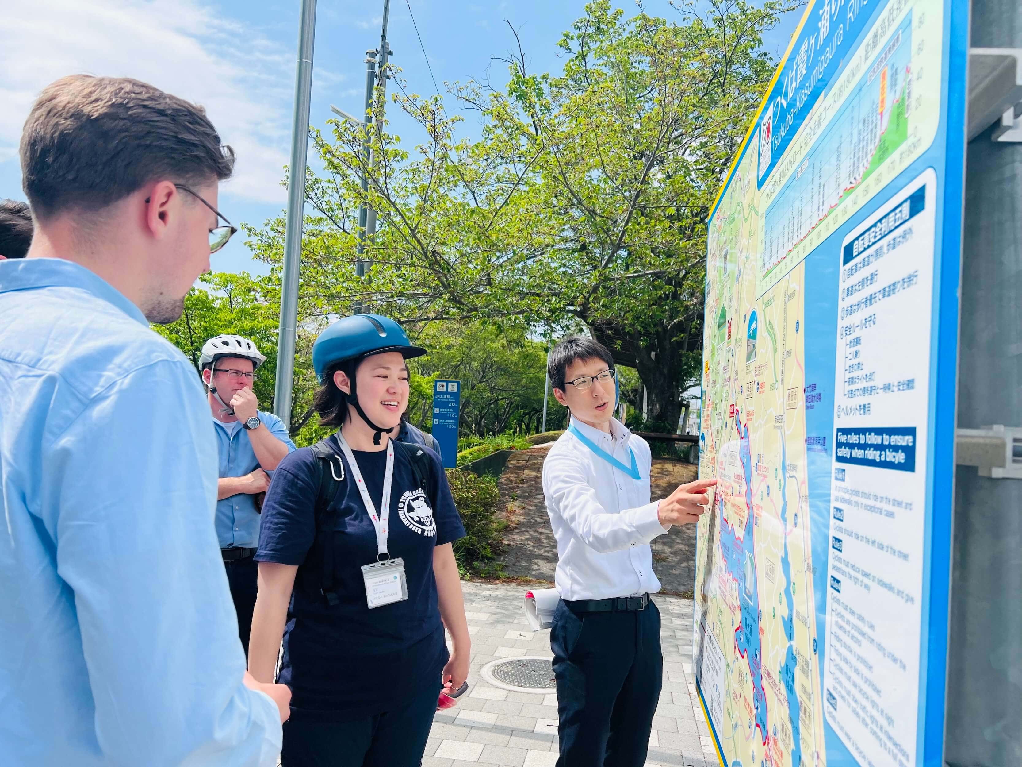 Gruppe diskutiert vor einer Infotafel zur Radroute in Tsukuba bei sonnigem Wetter