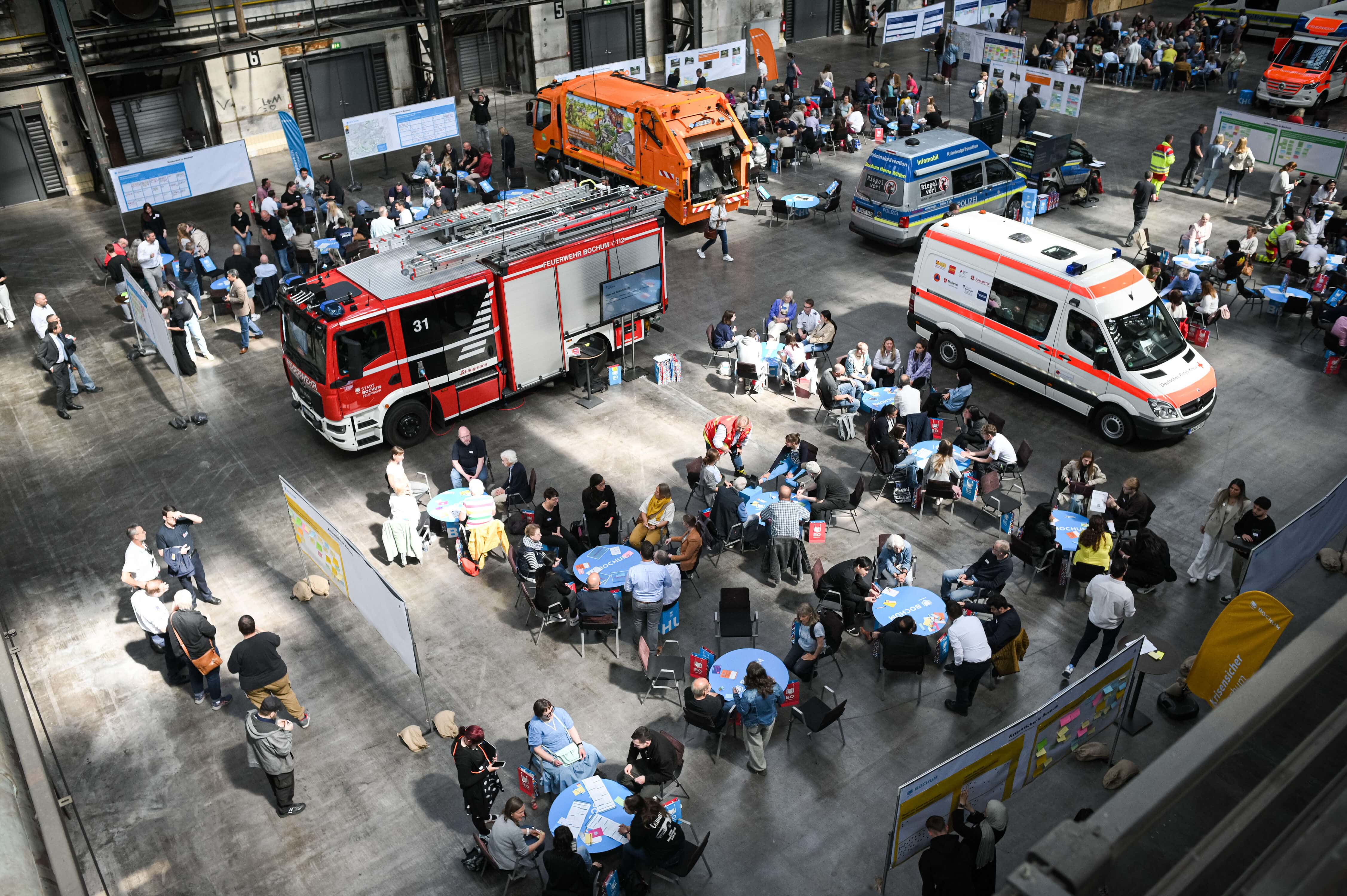 Blick in die Jahrhunderthalle mit Feuerwehr-, Rettungs- und Entsorgungsfahrzeugen sowie Diskussionsgruppen bei der Bürgerkonferenz 2025.
