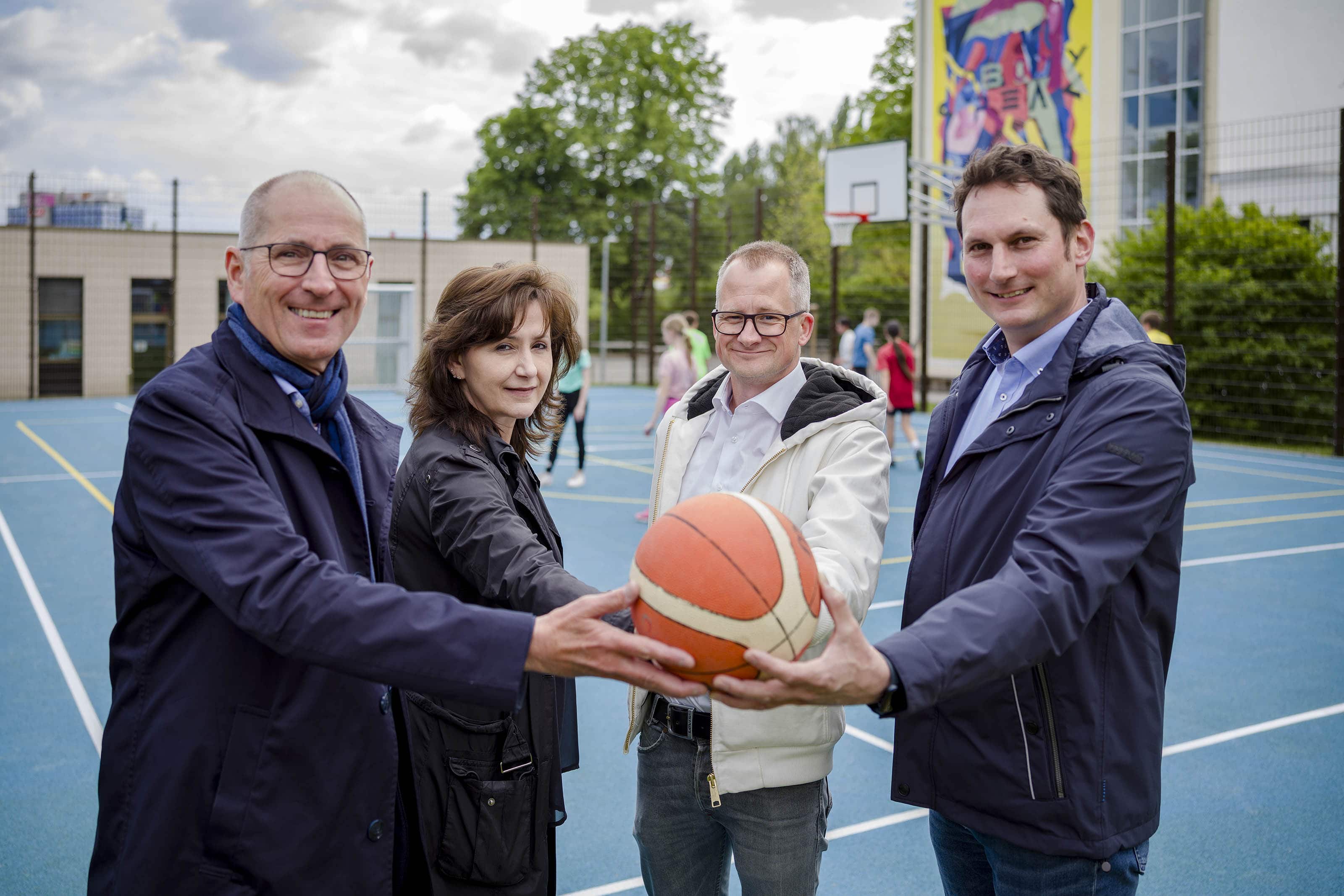 Vier Personen (v.l. Dietmar Dieckmann, Zoi Stefanou, Falko Hildebrandt und Marc Westerhoff) bei der Eröffnung des neuen Kleinspielfelds an der Pestalozzi-Realschule in Bochum-Wattenscheid, gemeinsam mit einem Basketball auf dem Spielfeld.