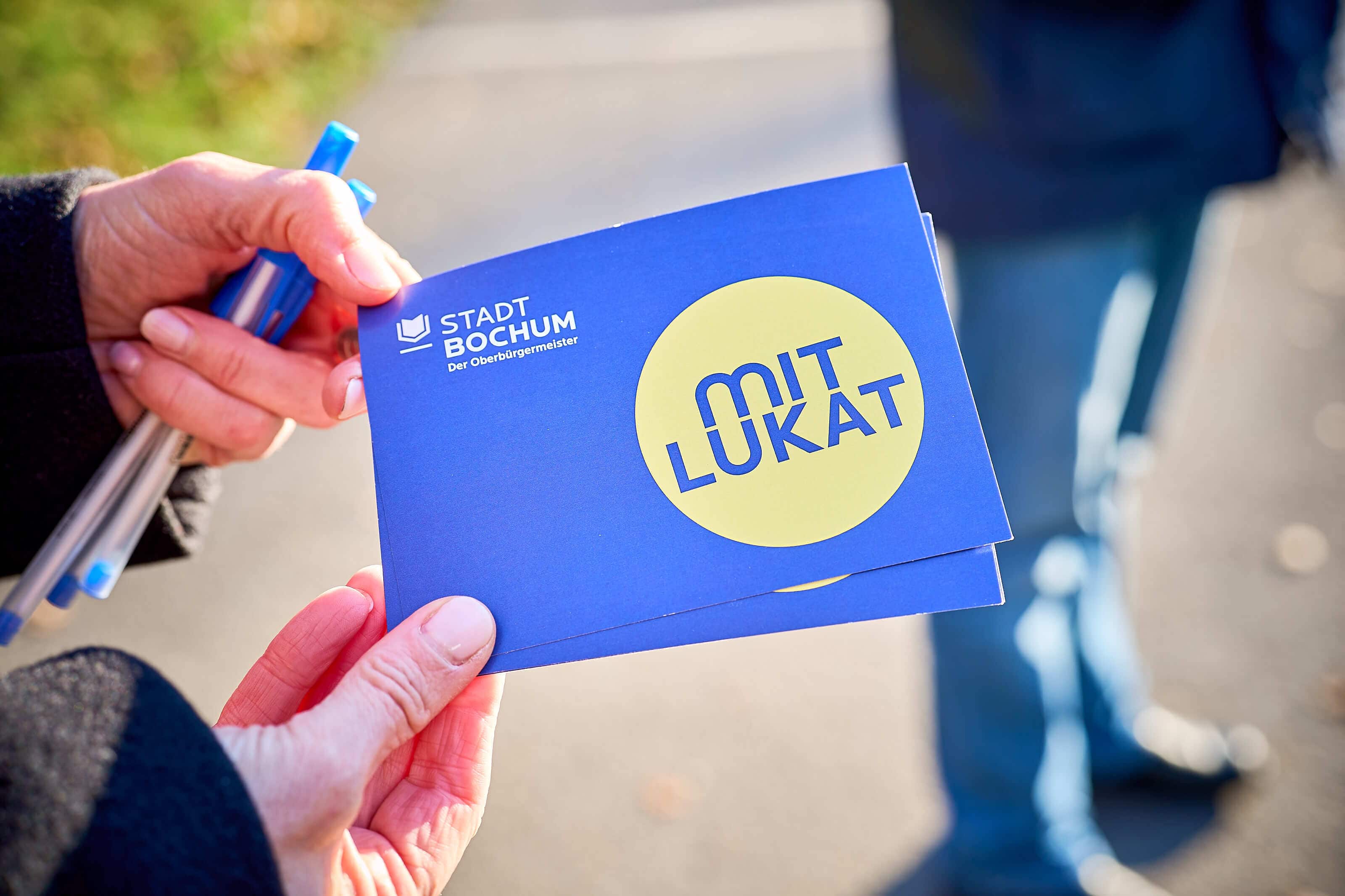 Zwei Hände halten zwei blaue Karten mit dem Aufdruck „mit Lukat“ und dem Stadtlogo von Bochum.